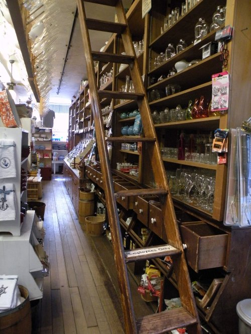 A wooden ladder leans against shelves filled with various glassware and kitchen items in a cozy store interior.