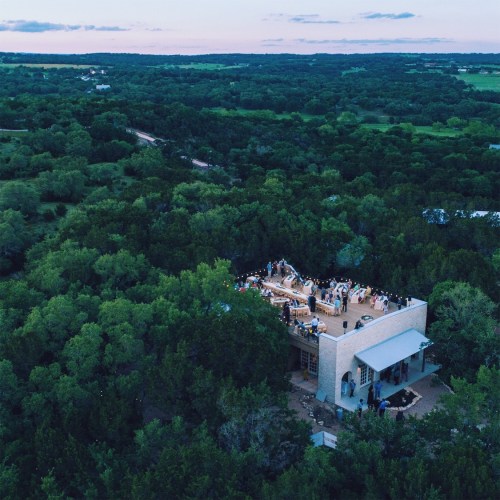 Aerial view of a gathering at a modern building surrounded by lush greenery and open fields at sunset.