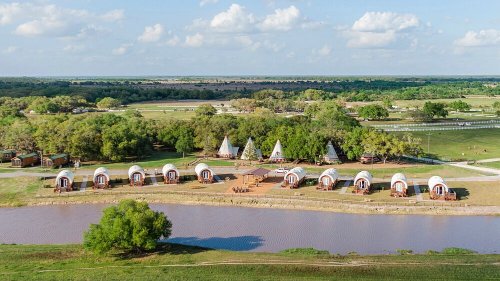 Aerial view of a campsite with teepees and wagons near a pond, surrounded by greenery and open fields.