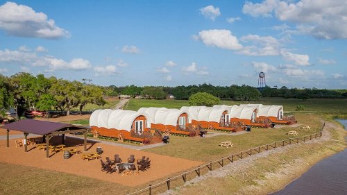 A scenic view of glamping tents arranged in a grassy area, with a shaded seating area and trees in the background.