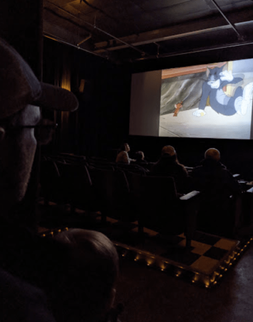 A person watches a cartoon on a big screen in a dimly lit theater, with a few other audience members visible.