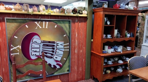 A cozy café interior featuring a whimsical clock sign and a wooden display shelf with various teacups and dishes.