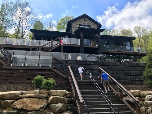 A modern house with a stone facade and large deck, surrounded by trees, with people on the deck and stairs.