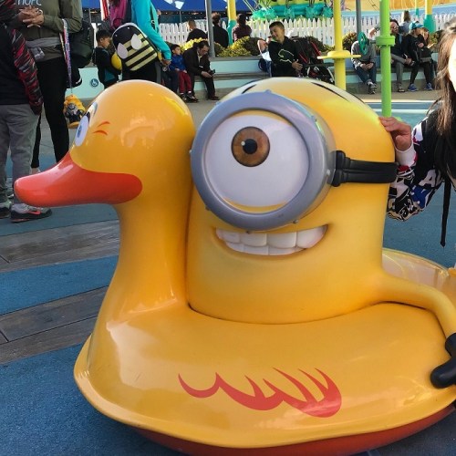 A child poses next to a large, cartoonish yellow duck with a single eye and a smiling face at a funfair.