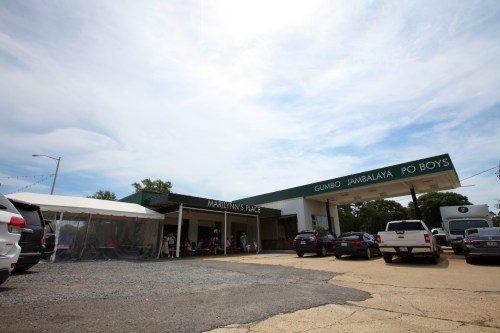 A restaurant building with a green awning, outdoor seating area, and cars parked in front under a cloudy sky.