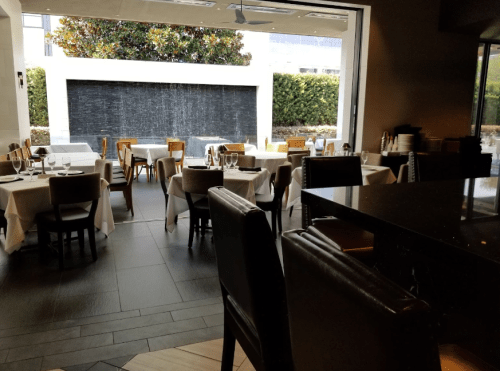 A modern restaurant interior with white tablecloths, wooden chairs, and a water feature in the background.