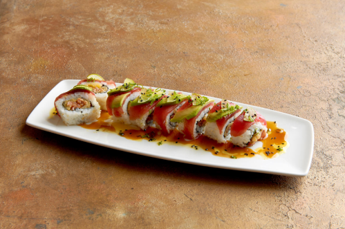 A sushi roll topped with avocado and tuna, drizzled with sauce, served on a white plate against a textured background.
