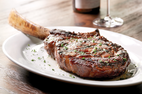 A perfectly grilled tomahawk steak garnished with herbs, served on a white plate with a glass of wine in the background.