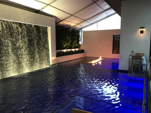A serene indoor space featuring a water wall, illuminated pool, and a fire feature, all under a tented ceiling.