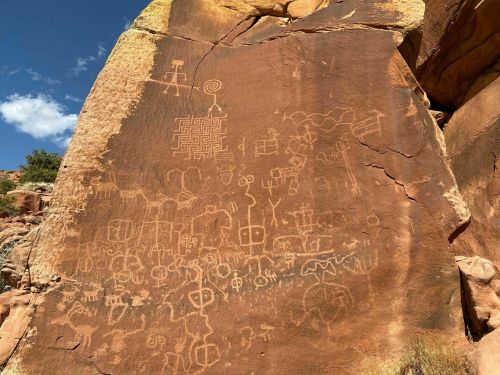 Petroglyphs etched into a large rock surface, featuring various abstract designs and symbols against a blue sky.