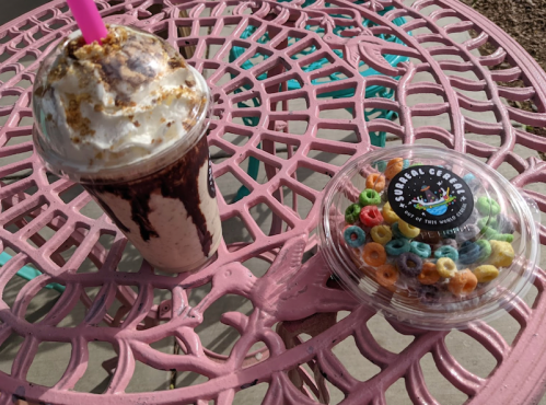 A milkshake topped with whipped cream and chocolate drizzle next to a container of colorful cereal on a pink table.