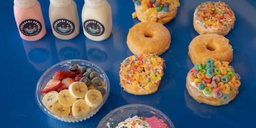 A colorful spread of donuts topped with cereal, a fruit bowl with strawberries and bananas, and milk bottles.