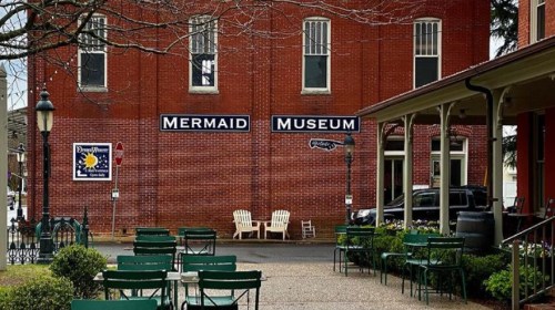Exterior of the Mermaid Museum, featuring a red brick building and outdoor seating area with green chairs.