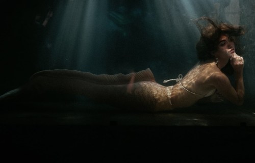 A woman in a shimmering mermaid costume poses underwater, surrounded by soft light and shadows.