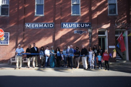 A group of people stands outside the Mermaid Museum, holding a ribbon for a grand opening ceremony.