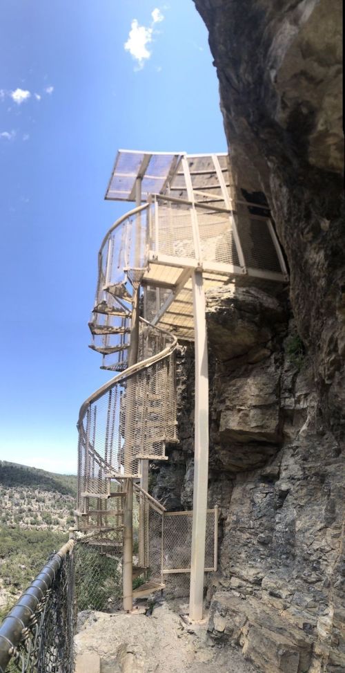 A spiral staircase attached to a rocky cliff, leading to a scenic overlook with blue skies in the background.