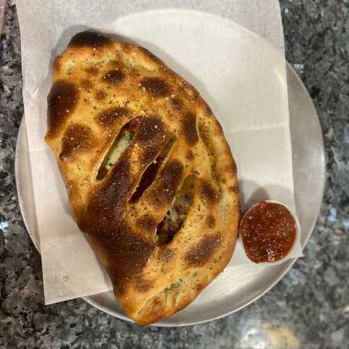 A golden-brown calzone on a plate, served with a small cup of marinara sauce on the side.