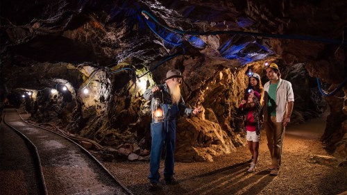 A tour guide with a lantern shows a couple around a dimly lit mine, surrounded by rocky walls and soft lighting.