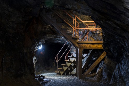 Dimly lit underground mine with wooden stairs and metal railings, surrounded by rocky walls and gravel floor.