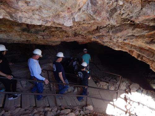 A group of people wearing hard hats descends a wooden staircase into a cave. Rocks and cave walls are visible.