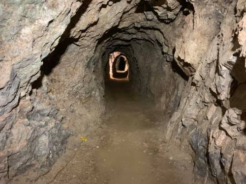 A dimly lit tunnel carved through rocky terrain, with a dirt floor and openings in the distance.