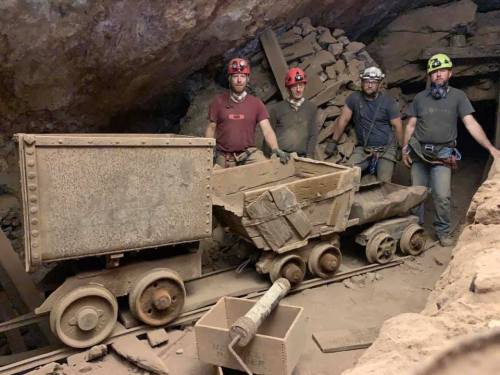 Four miners stand beside old mining carts in a dusty underground tunnel, wearing helmets and headlamps.