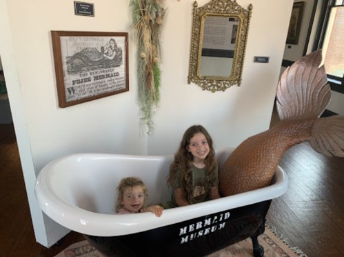 Two children sit in a vintage bathtub at the Mermaid Museum, one with a mermaid tail, surrounded by whimsical decor.