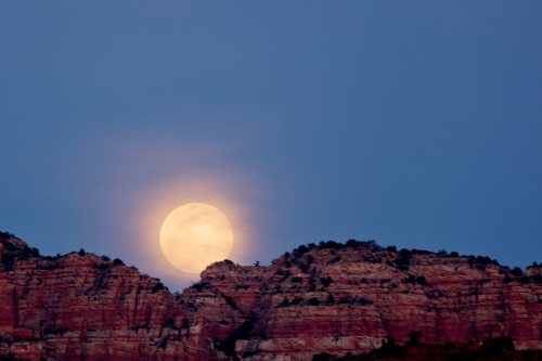 A large full moon rises behind rugged red rock formations under a twilight sky.