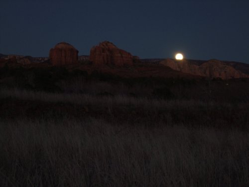 A moonrise over rocky formations, with tall grass in the foreground, set against a darkening sky.