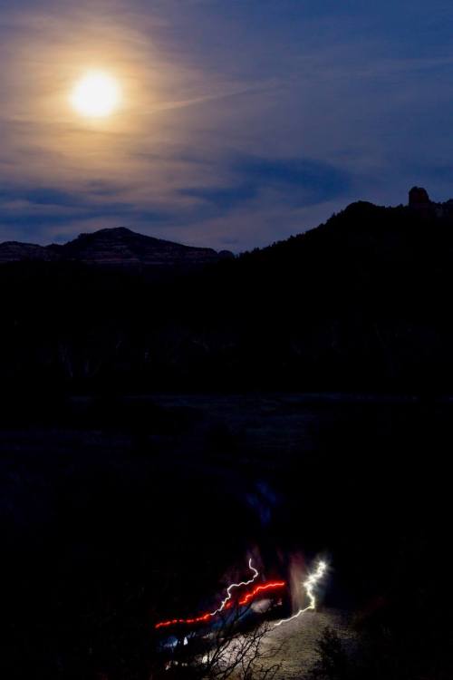 A moonlit landscape with a glowing moon, silhouetted mountains, and streaks of light from a vehicle below.