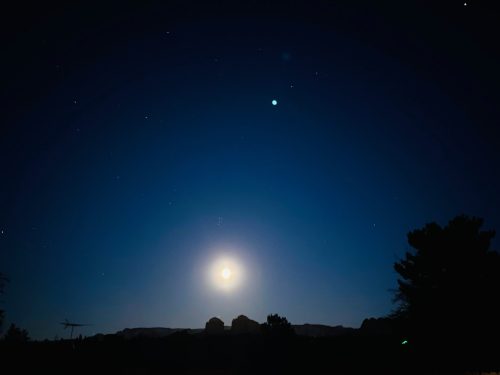 A serene night sky with a bright moon, stars, and a planet visible above a dark silhouette of trees and mountains.