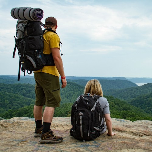 Two hikers with backpacks stand on a rocky ledge, overlooking a lush green valley and distant hills.