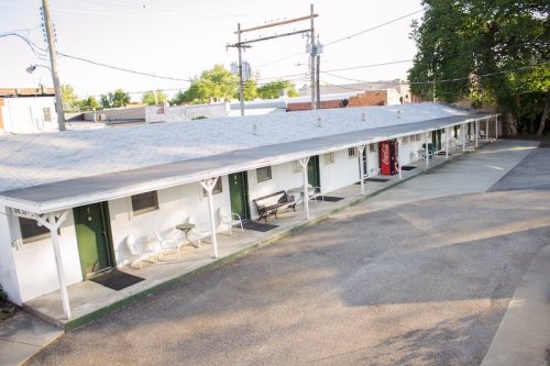 A row of simple motel rooms with a gray roof, green doors, and a vending machine in a quiet outdoor setting.