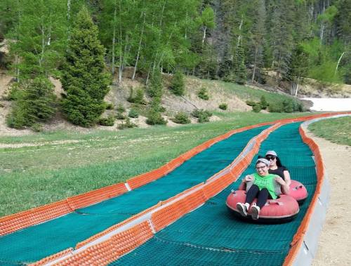 Two people enjoy tubing down a grassy slope, surrounded by trees and a clear blue sky.
