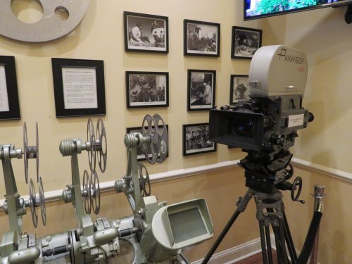 A vintage film projector and camera displayed in a museum, with black-and-white photos on the wall behind them.