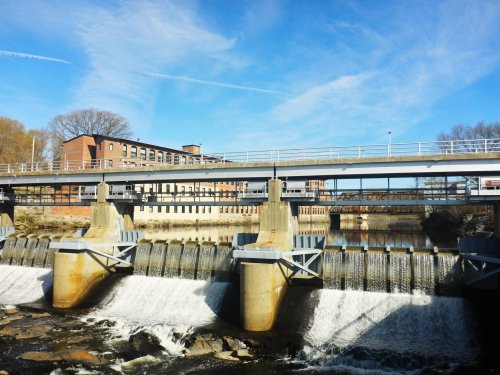 A dam with flowing water beneath a bridge, with buildings and trees in the background under a clear blue sky.