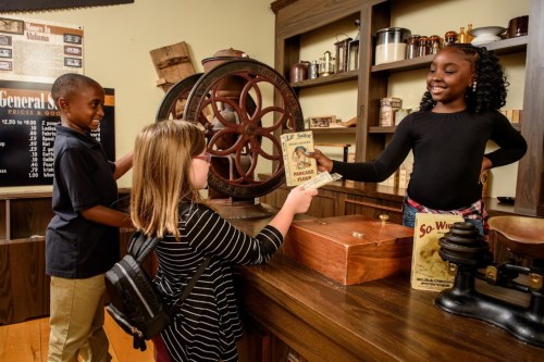 Children interacting in a vintage general store, examining old-fashioned items and sharing a flyer.