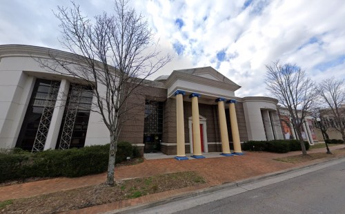 A modern building with columns and large windows, surrounded by trees and a brick walkway under a cloudy sky.