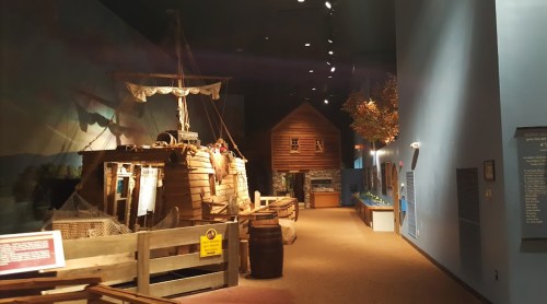 A museum exhibit featuring a wooden ship, a rustic house, and various displays in a dimly lit hallway.