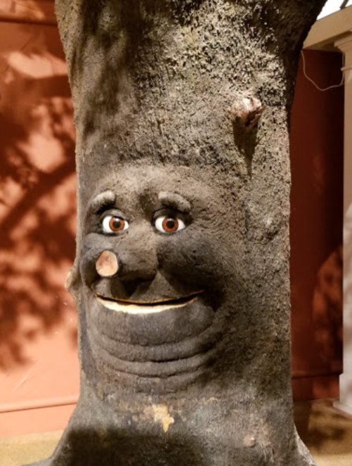 A tree trunk with a carved face, featuring eyes, a nose, and a smiling mouth, set against a reddish background.