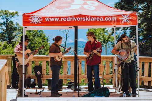 A band performs under a red tent in a scenic outdoor setting, surrounded by trees and mountains.