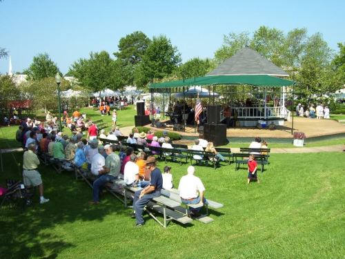 A lively outdoor event with a band performing on stage, surrounded by an audience seated on benches in a park.