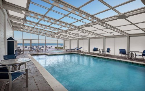 Indoor swimming pool with lounge chairs and tables, surrounded by large windows overlooking a scenic view.