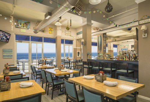 Bright restaurant interior with wooden tables, blue chairs, and ocean views through large windows. Decorated with beach-themed art.