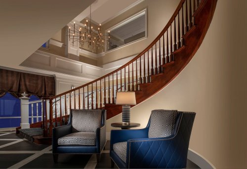 Elegant interior featuring a curved staircase, two blue chairs, a side table, and a chandelier above.