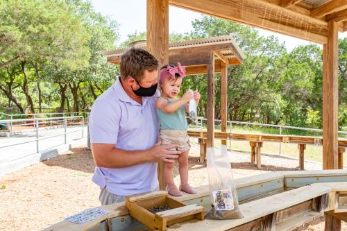 A man in a mask helps a toddler with a craft activity at an outdoor park. Trees and a wooden structure are in the background.