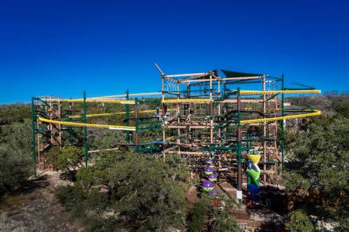 A large outdoor adventure park structure with slides and climbing features, surrounded by trees under a clear blue sky.