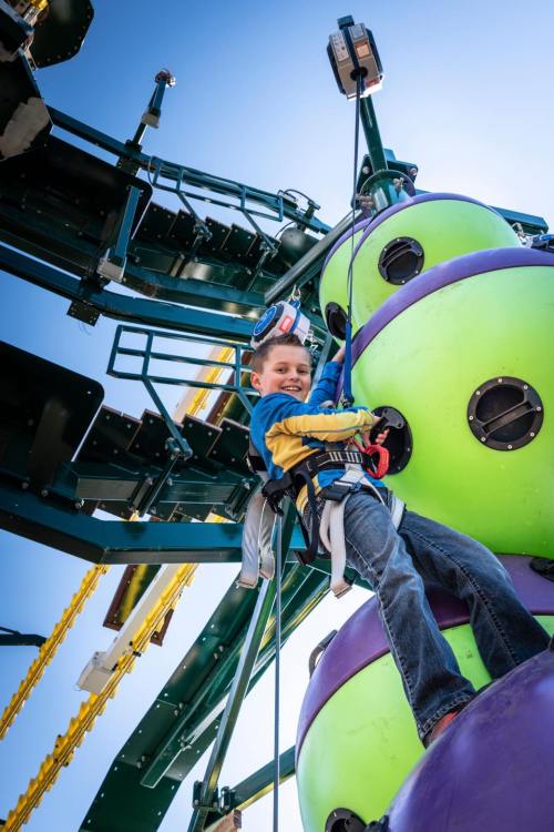 A smiling child climbs a colorful structure, secured with a harness and rope, against a clear blue sky.