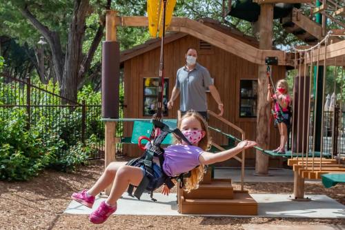 A girl in a mask swings on a zip line while an adult supervises nearby, with another child on a climbing structure.