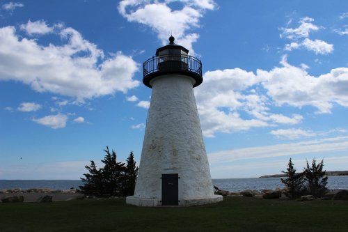 A white lighthouse stands by the ocean under a blue sky with clouds, surrounded by green grass and trees.